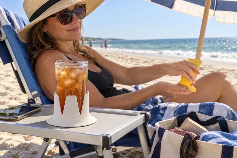 Adult reclining in low beach chair under umbrella with open reusable plastic iced-tea cup in Steadi on wide flat beach-chair side tray designed for chair and fully supporting base. AI illustration.